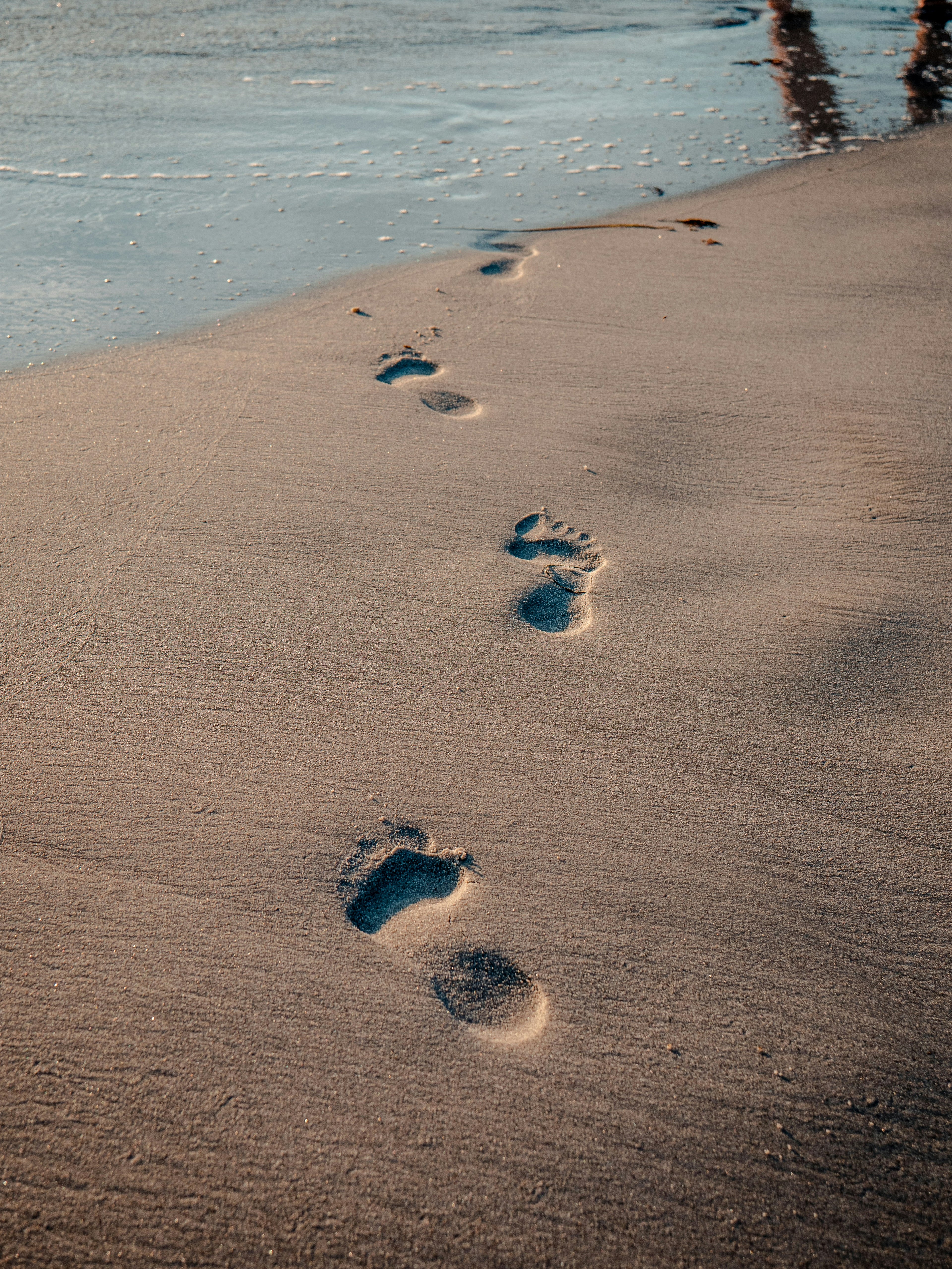 Fußabdrücke im Sand eines Strandes, die sich sanft in Richtung Wasser ziehen, mit einem kleinen Teil des Meeres im Hintergrund, der sanft gegen den Strand schlägt.