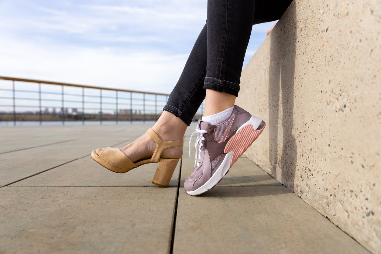 Frau mit einem Stöckelschuh und einem Sneaker an den Füßen, die unterschiedliche Schuhstile kombiniert.