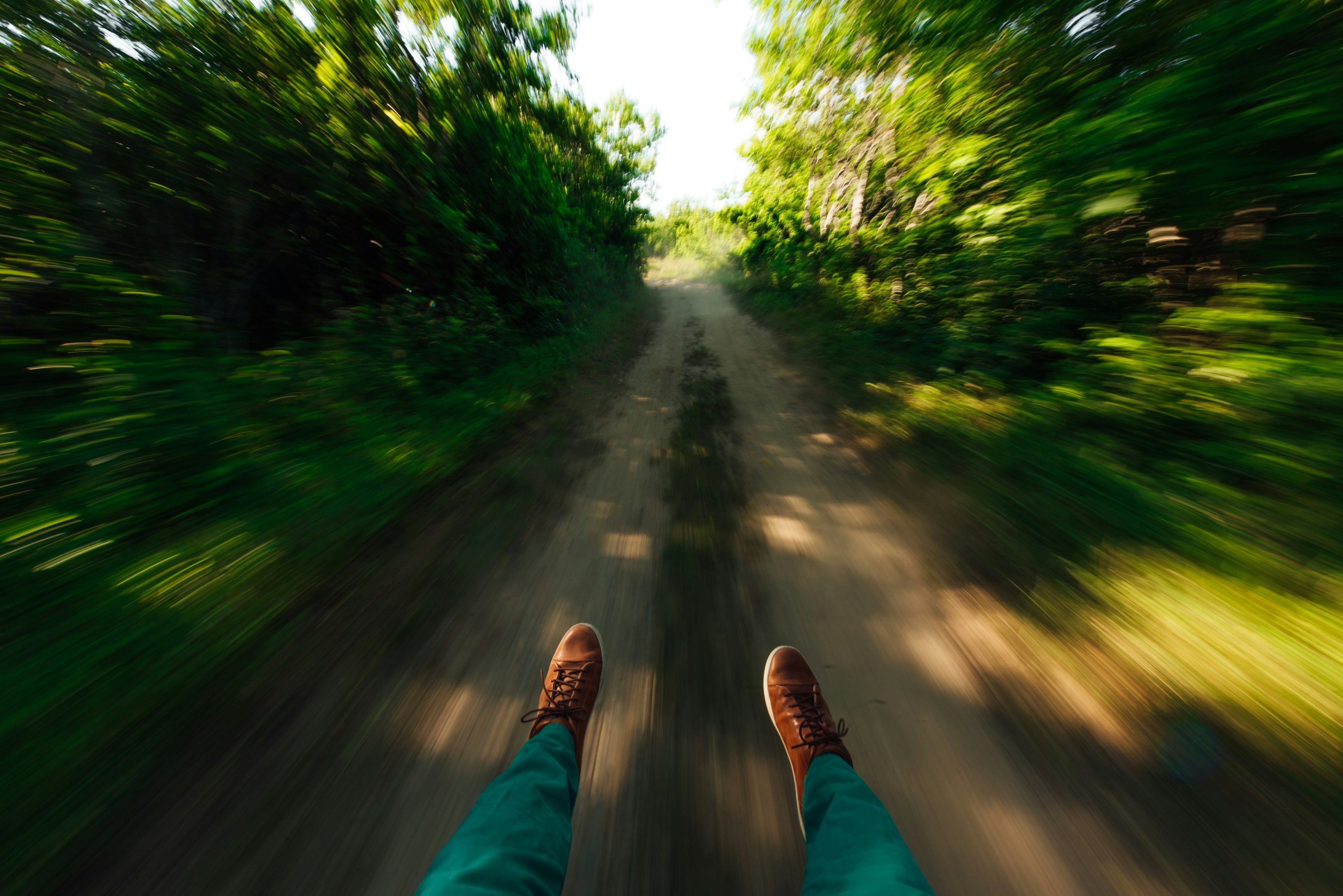 Foto zeigt aus der Perspektive einer Person, die auf einem Weg durch einen grünen, dicht bewachsenen Wald fährt. Die Beine und Füße der Person sind sichtbar, während der Hintergrund in Bewegung unscharf wirkt, was ein Gefühl von Geschwindigkeit vermittelt.