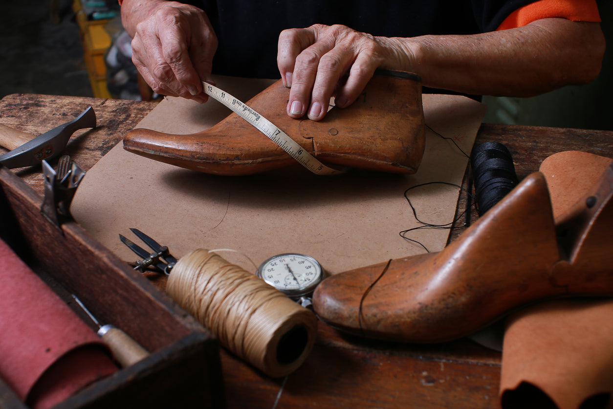 Ein Handwerker misst sorgfältig einen Leisten mit einem Maßband. Im Hintergrund sind verschiedene Werkzeuge und Materialien sichtbar, die in der Schuhherstellung verwendet werden, was die handwerkliche Umgebung verdeutlicht.