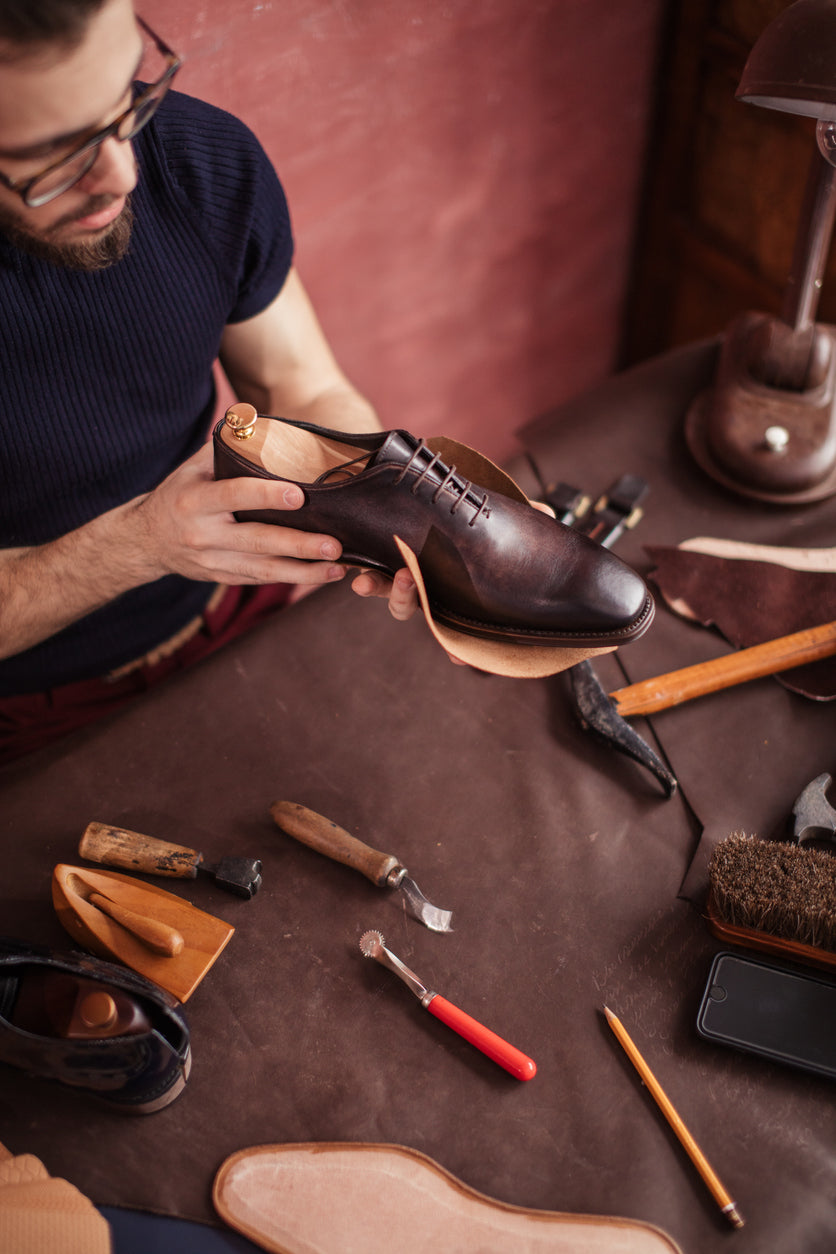 Ein Mann hält einen eleganten Maßschuh in der Hand und kontrolliert ihn sorgfältig auf Qualität und Details. Sein Gesichtsausdruck zeigt Konzentration und Zufriedenheit, während er die Verarbeitung des Schuhs überprüft.