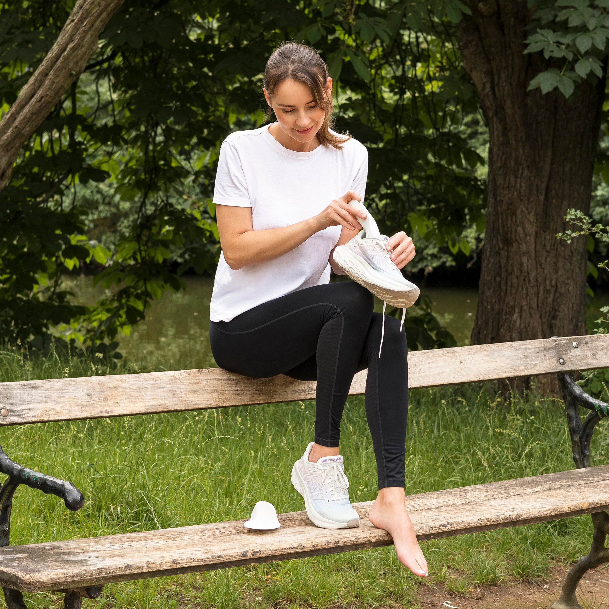 Eine Frau sitzt auf einem Holzzaun und legt eine orthopädische Einlage in ihren Sportschuh. Im Hintergrund erstreckt sich eine grüne Wiese, die eine frische und natürliche Atmosphäre schafft. Die Frau wirkt entspannt und konzentriert bei ihrer Tätigkeit.