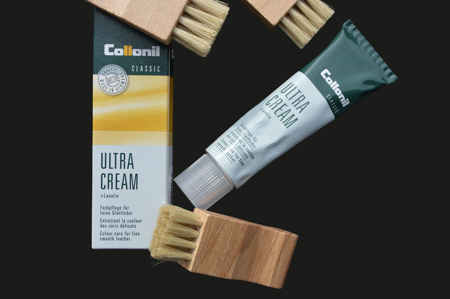 Su uno sfondo scuro, una tubetto di Collonil Ultra Cream, il packaging della crema e tre spazzole per l'applicazione sono disposti. I prodotti sono chiaramente visibili e si distinguono nettamente dallo sfondo, evidenziando i loro dettagli e il design.